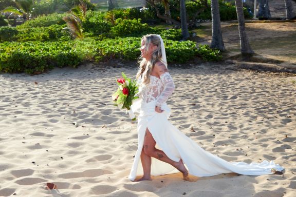 Sandy Beach Wedding Ceremony Bride in a flowing white dress walks on the beach holding a bouquet.