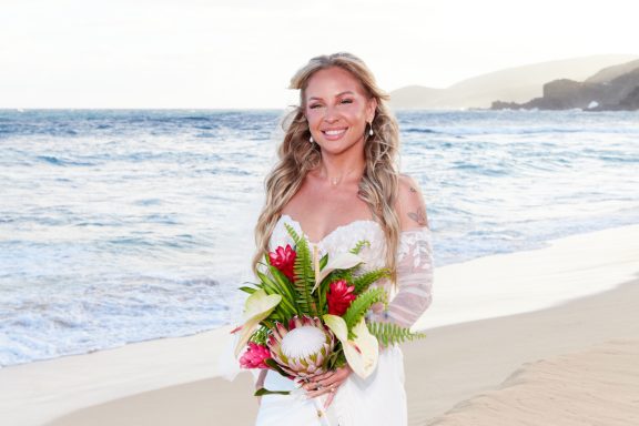Sandy Beach Wedding Ceremony Smiling woman in a wedding dress holding a bouquet, standing by the beach.