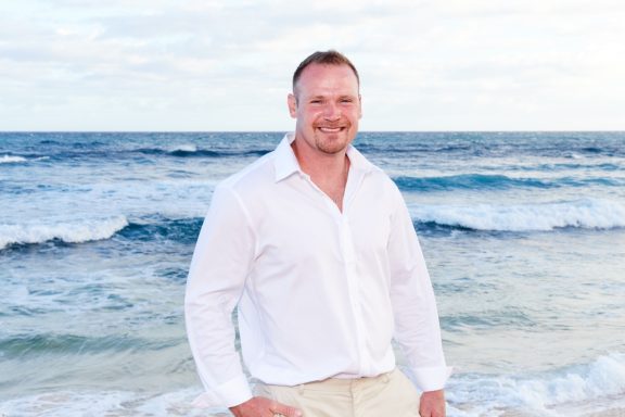 Sandy Beach Wedding Ceremony Smiling man in a white shirt standing by the beach with ocean waves in the background.