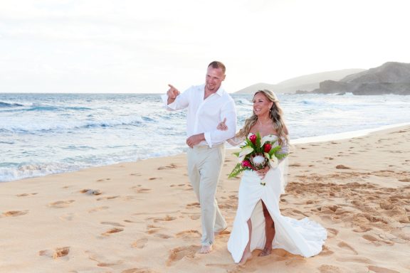 Sandy Beach Wedding Ceremony Couple walking along a beach, bride holding a bouquet, smiling.