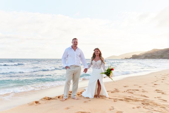 Sandy Beach Wedding Ceremony A couple in wedding attire holds hands on a sandy beach with ocean waves in the background.