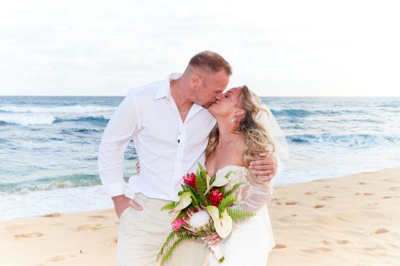 Sandy Beach Wedding Ceremony Couple kissing on a beach, holding a bouquet, with ocean waves in the background.