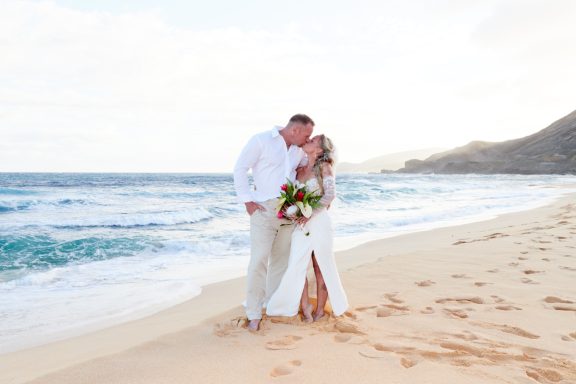Sandy Beach Wedding Ceremony Couple in white attire sharing a kiss on a beach, with waves and mountains in the background.