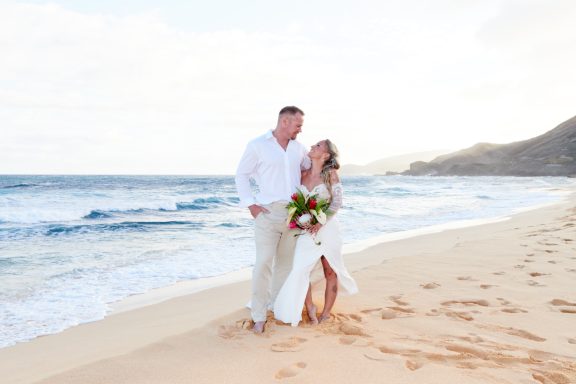 Sandy Beach Wedding Ceremony Couple standing on the beach, smiling, with waves and a sunset in the background.