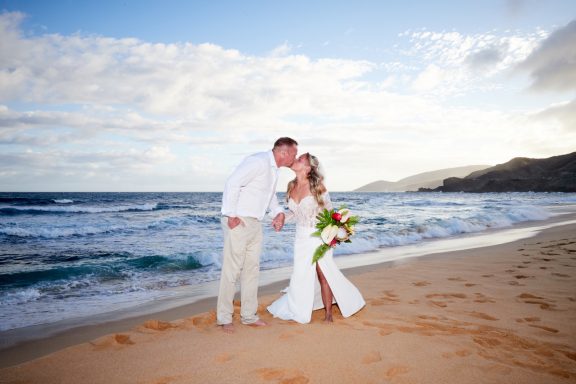 Sandy Beach Wedding Ceremony Couple in wedding attire kissing on a beach at sunset with waves in the background.