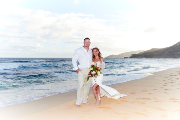 Sandy Beach Wedding Ceremony Couple in white clothing standing on a beach, holding a bouquet and smiling.