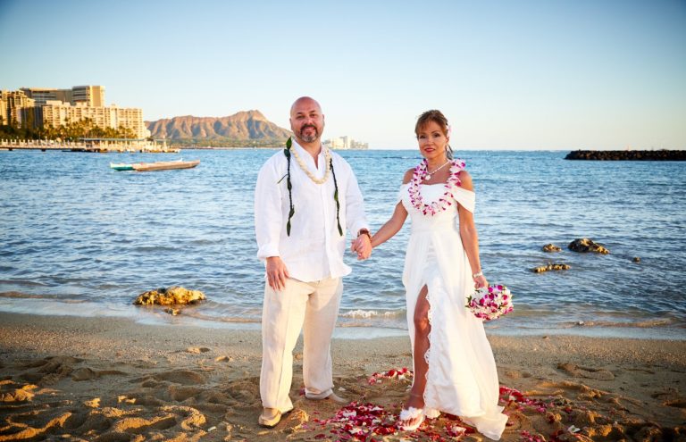 Couple in wedding attire holding hands on a beach with ocean and Waikiki in the background at Duke Kahanomoku Beach.