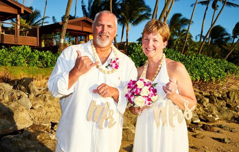 Couple in white attire holding floral bouquet on a beach with palm trees in the background at Portlock Beach.