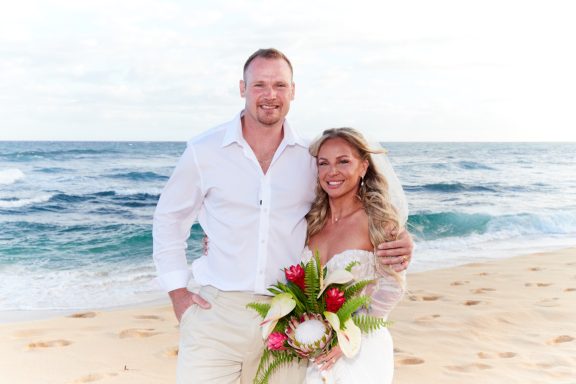 Sandy Beach Wedding Ceremony A couple in wedding attire stands on a beach holding tropical flowers.