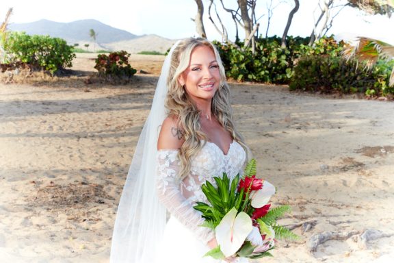 Sandy Beach Bride Bride in a white gown, holding a bouquet, smiling in a sandy outdoor setting.