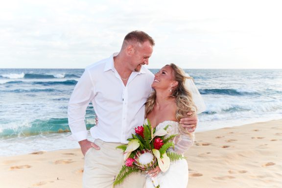 Sandy Beach Wedding Ceremony A couple gazes into each other's eyes on a beach, holding a bouquet and smiling.
