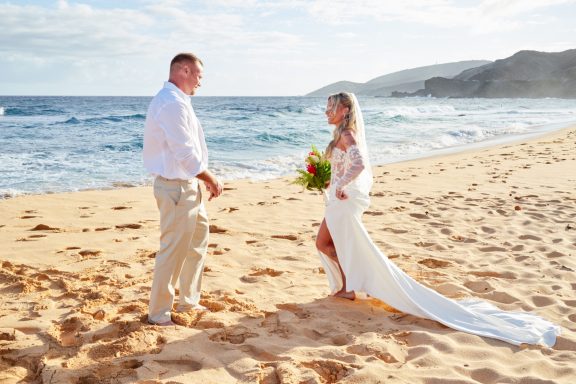 Sandy Beach Wedding Ceremony Couple in wedding attire on a beach at sunset, holding a bouquet and smiling.