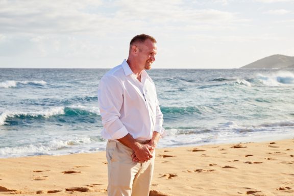 Sandy Beach Wedding Ceremony Man in a white shirt stands by the shore, with waves and sand in the background.