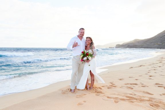 Sandy Beach Wedding Ceremony A couple in wedding attire smiles on a beach, holding a bouquet with ocean waves in the background.